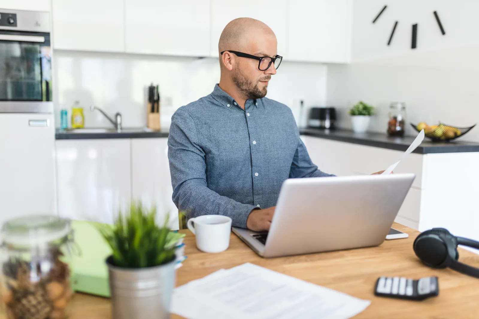 A man in glasses reads a document as he types on a laptop at his kitchen table, papers and a calculator nearby