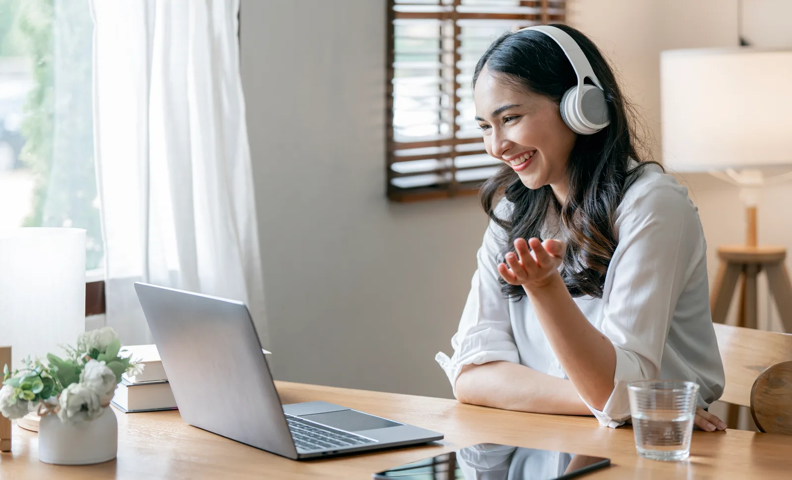 A smiling woman in white headphones talks to her laptop camera at a wooden desk beside a sunny window