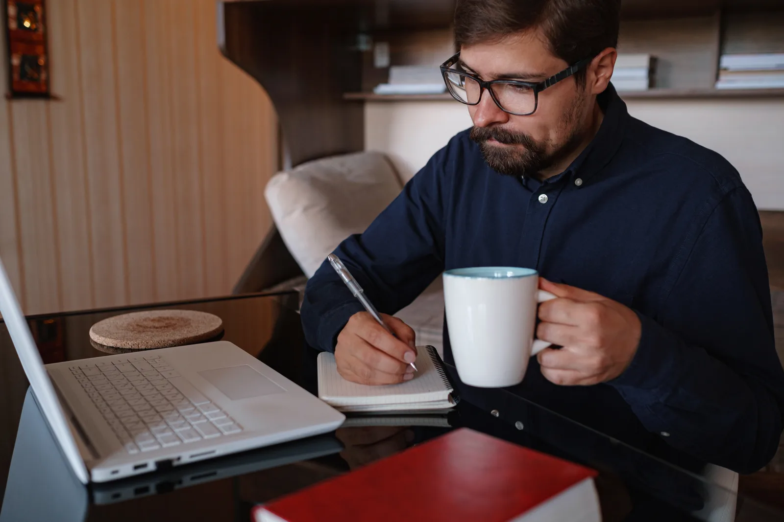 A man in glasses writes in a notepad beside his laptop at home, holding a mug of coffee in his other hand