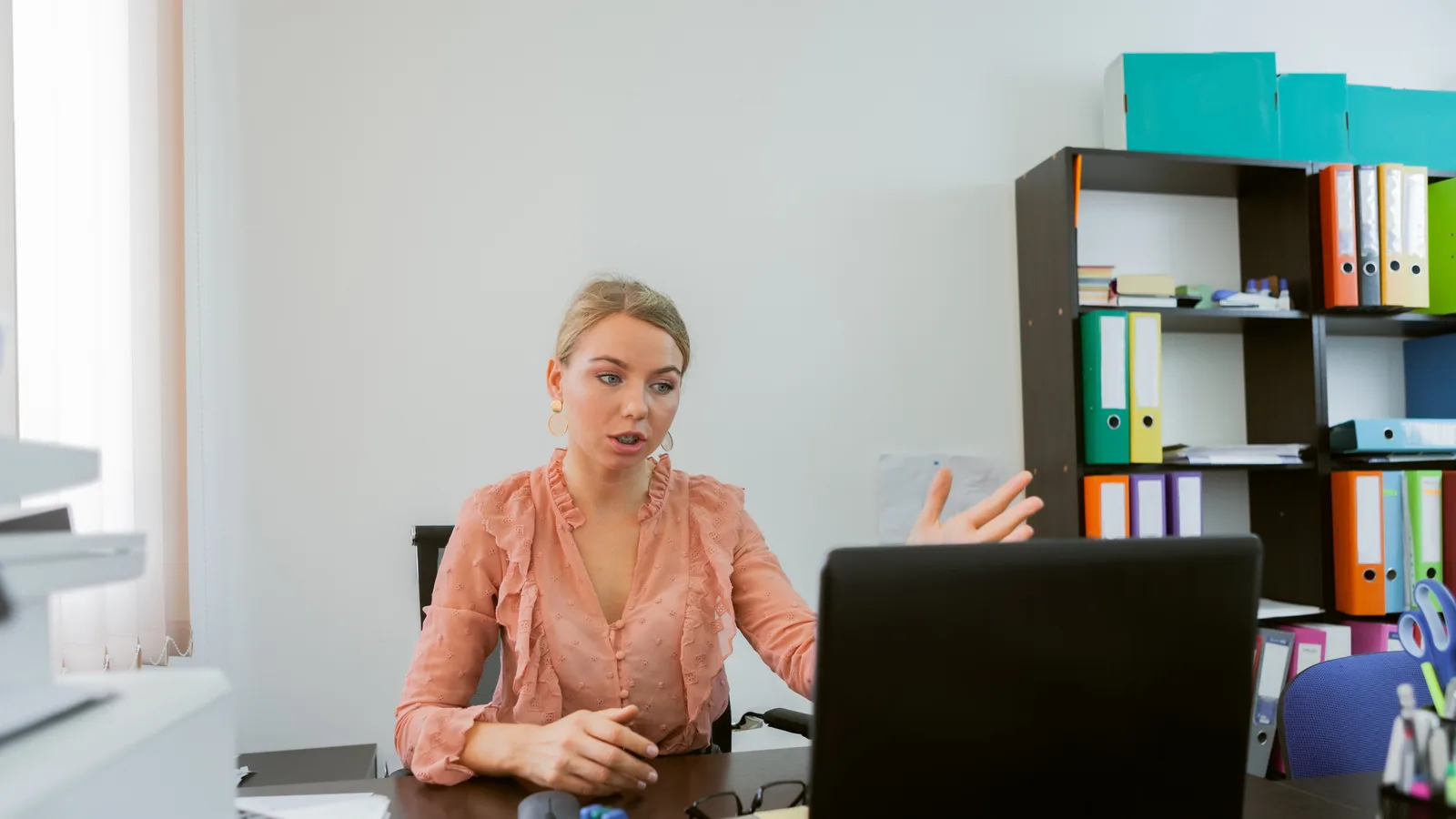 Account manager at a small agency office desk on a video call with a remote colleague shown on a laptop