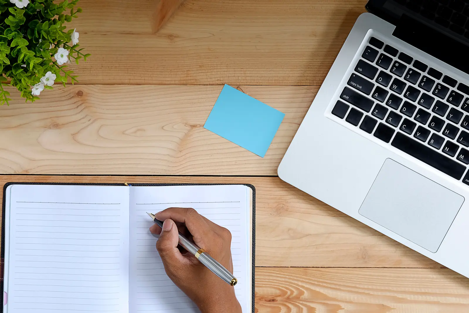 A hand ticks through a notebook checklist beside a laptop and a sticky note