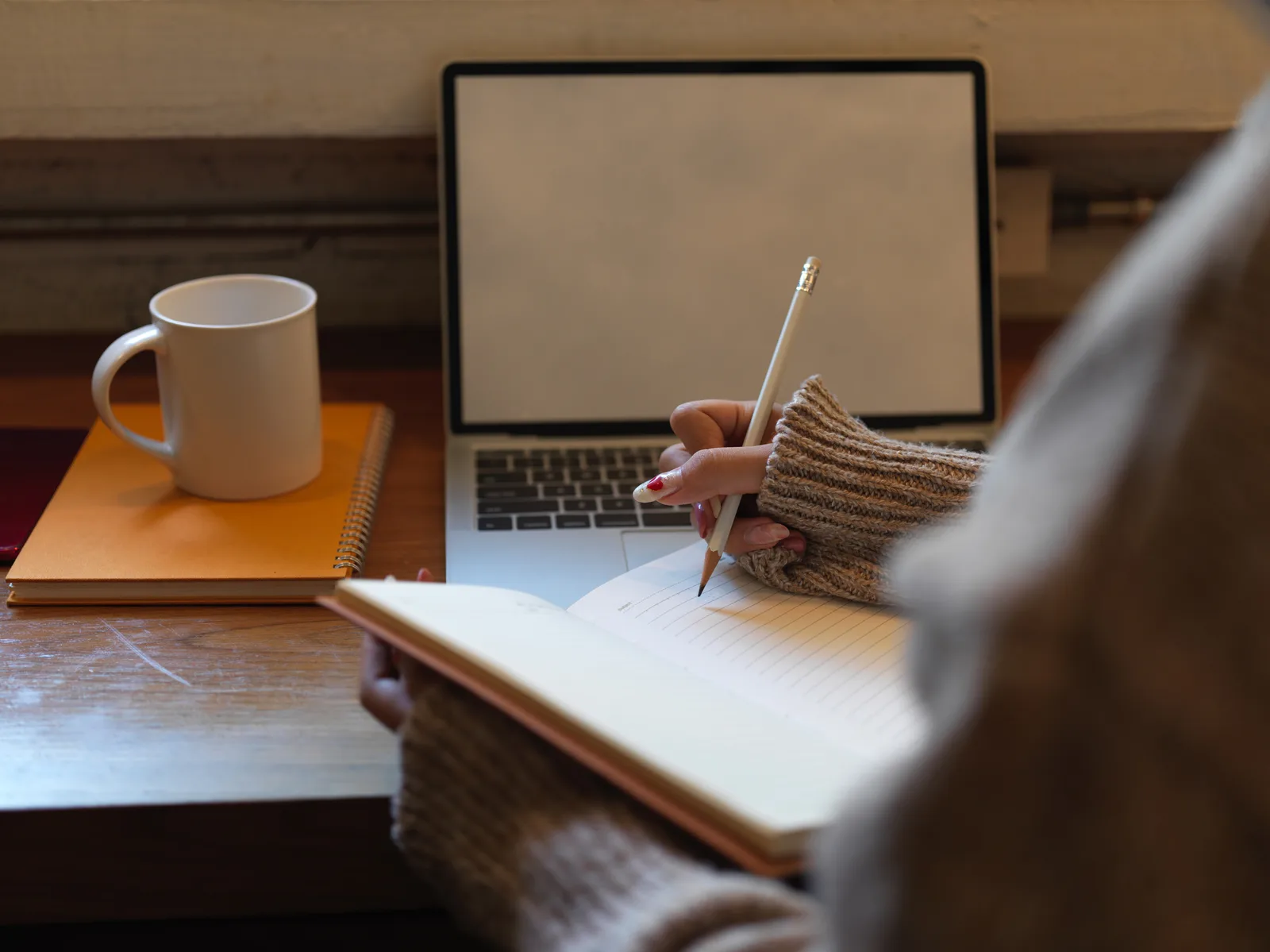 A hand writes in a notebook beside a laptop and a mug, working out the draft on paper first