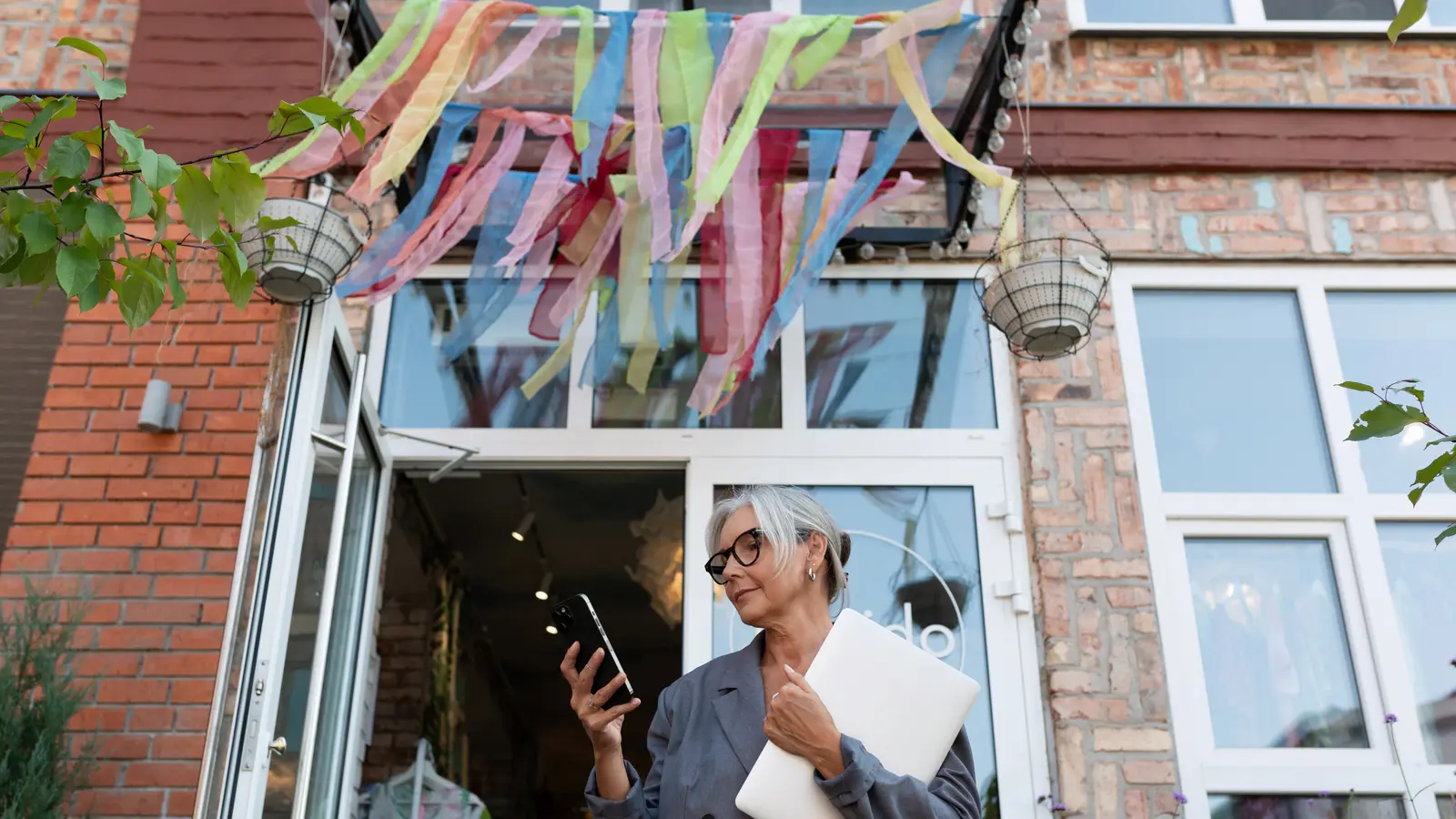 Shop owner in the doorway of her small store checking her phone with a laptop under her arm