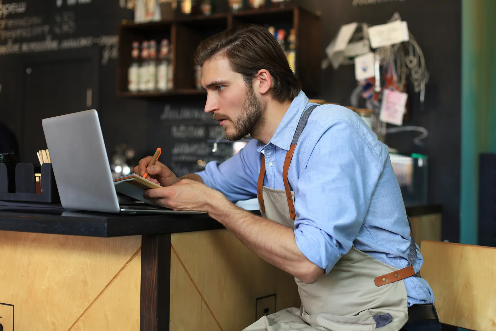 Cafe owner in an apron at his counter working on a laptop and taking notes