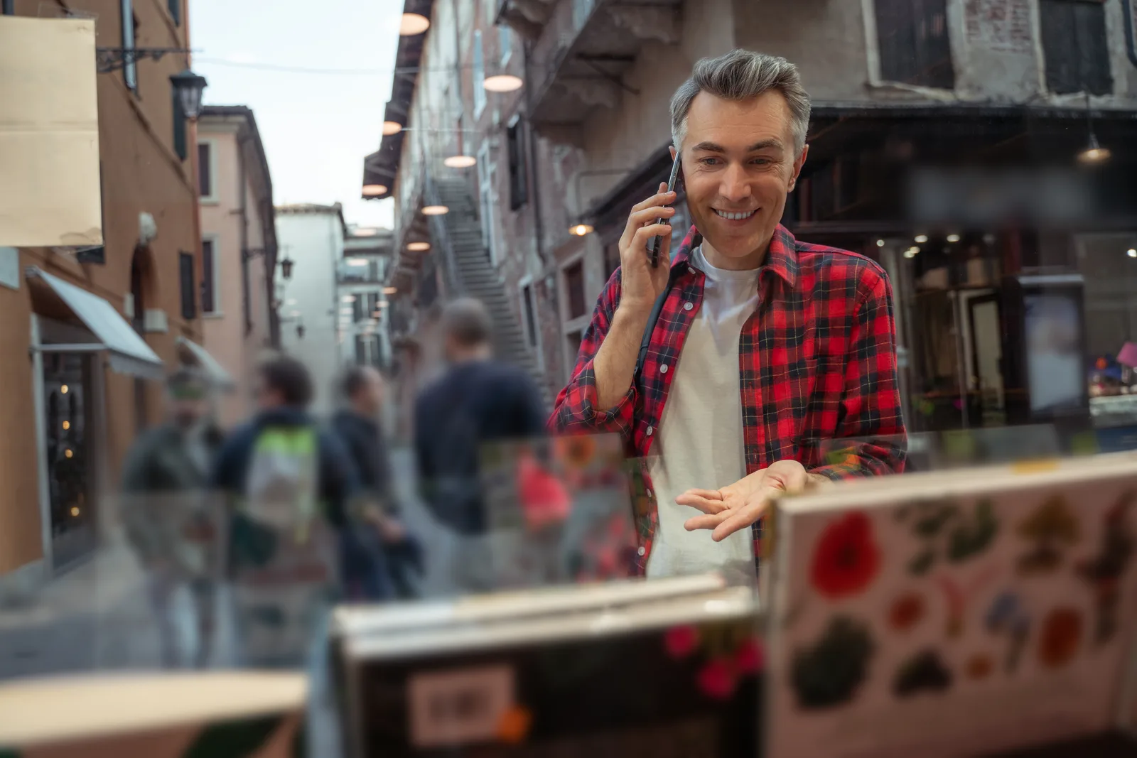 Shop owner in a plaid shirt taking a call at his storefront window on a narrow street
