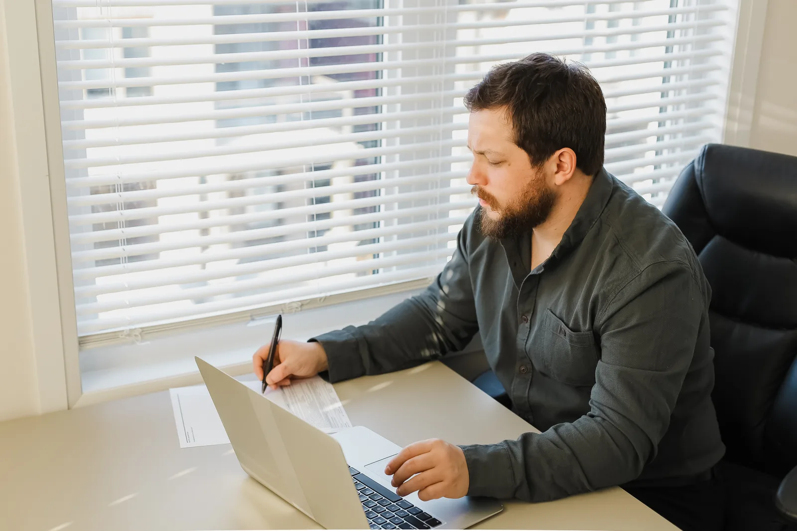 A bearded man at a laptop by a window writes notes on a printed sheet beside his keyboard