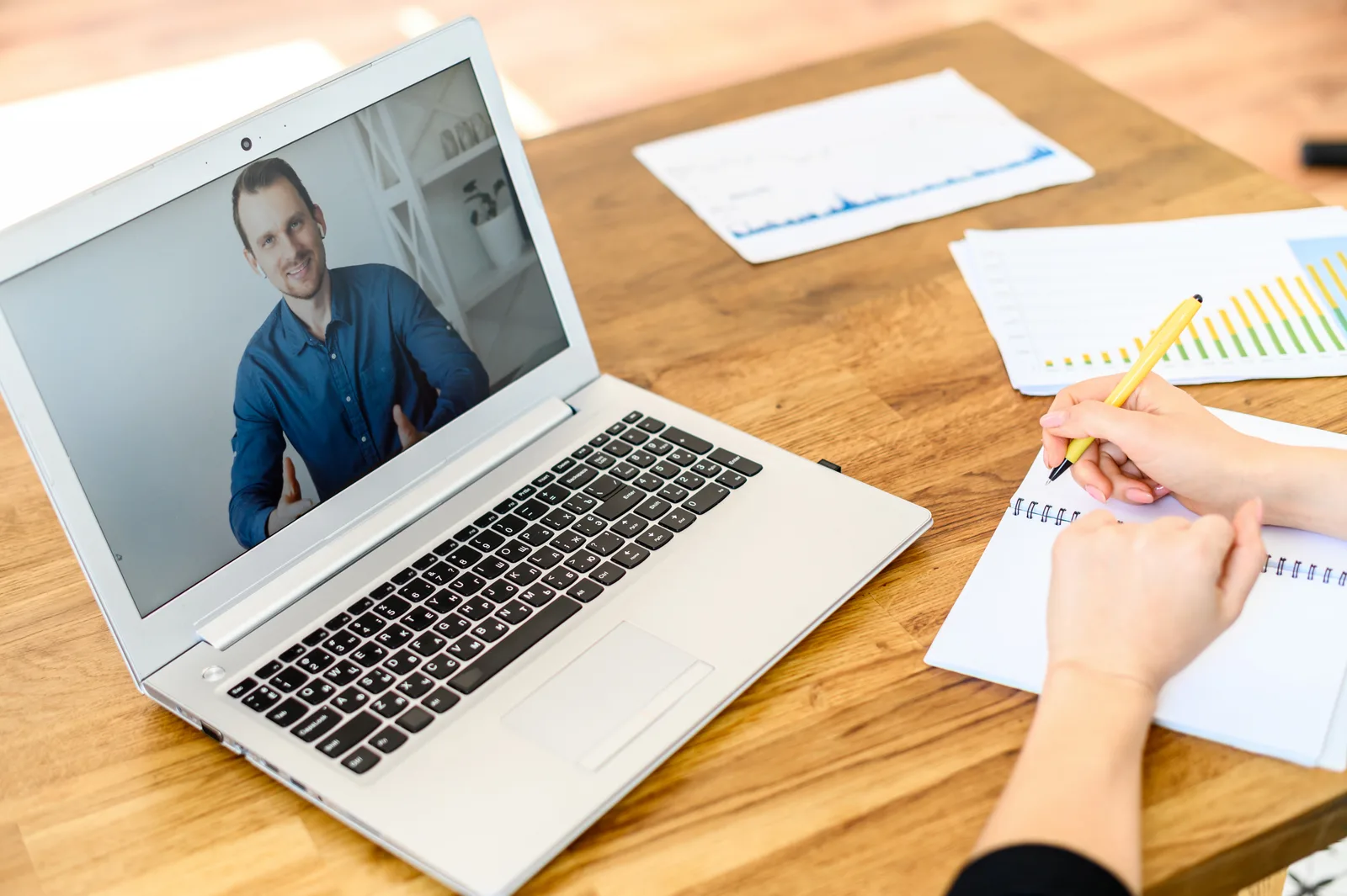 A man smiles from a laptop video call while the interviewer takes notes beside printed charts on the desk
