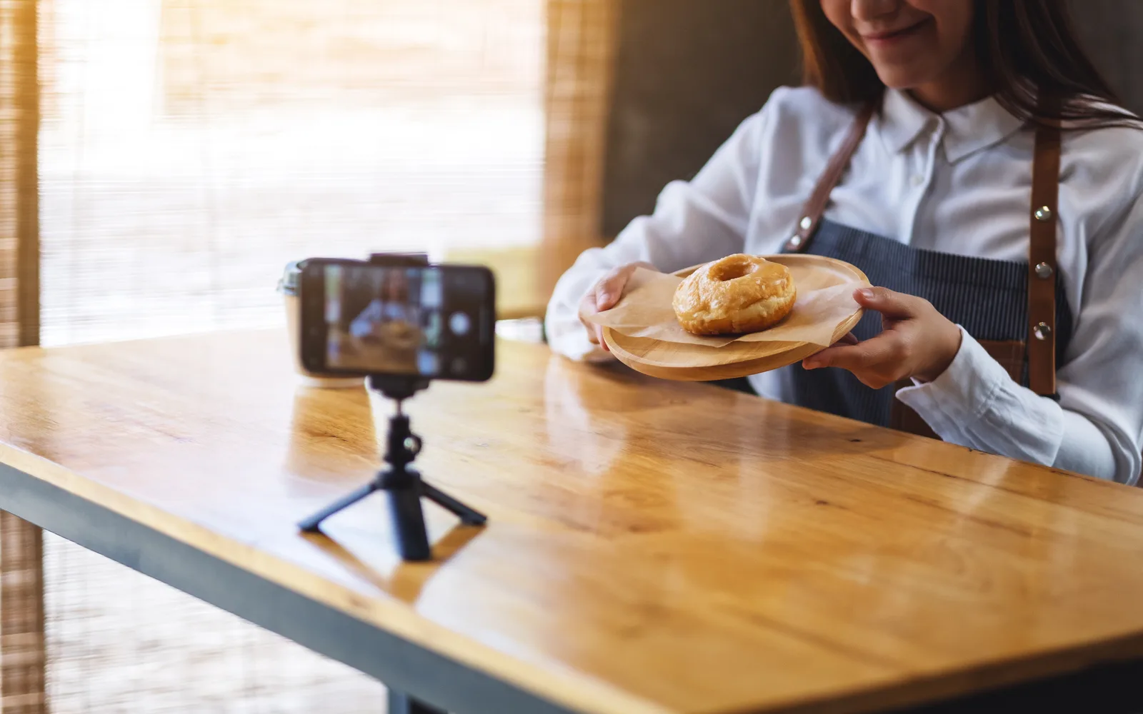 A woman in an apron films a donut on a plate with a phone on a small tripod at a wooden table