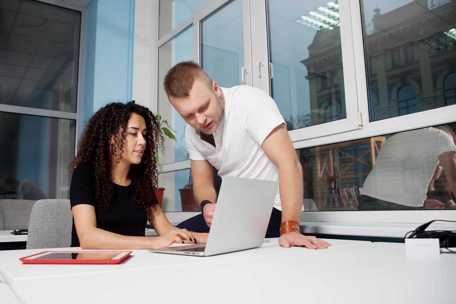 A man leans in to walk a colleague through something on her laptop in a bright, window-lined office