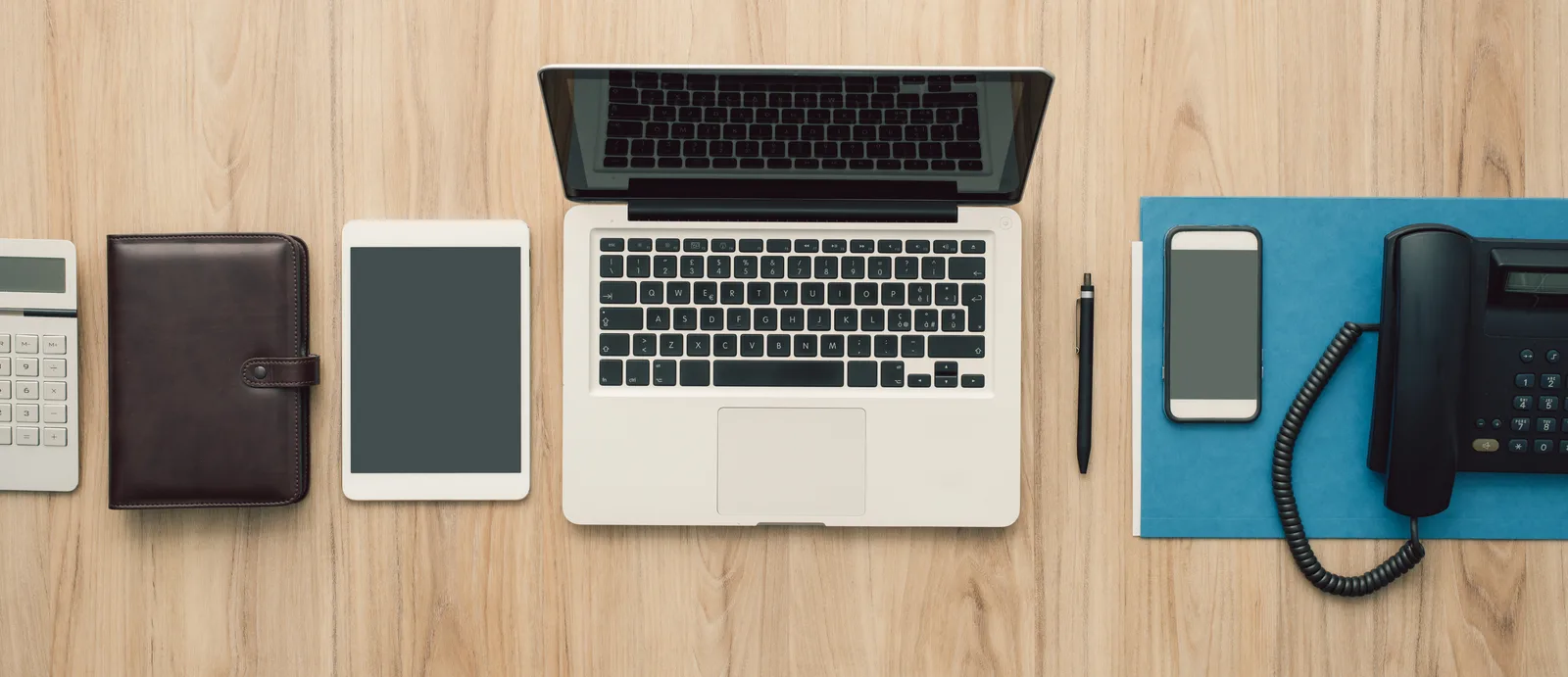 A tidy desk from above with a laptop, a tablet, a phone and a notebook laid out in a grid