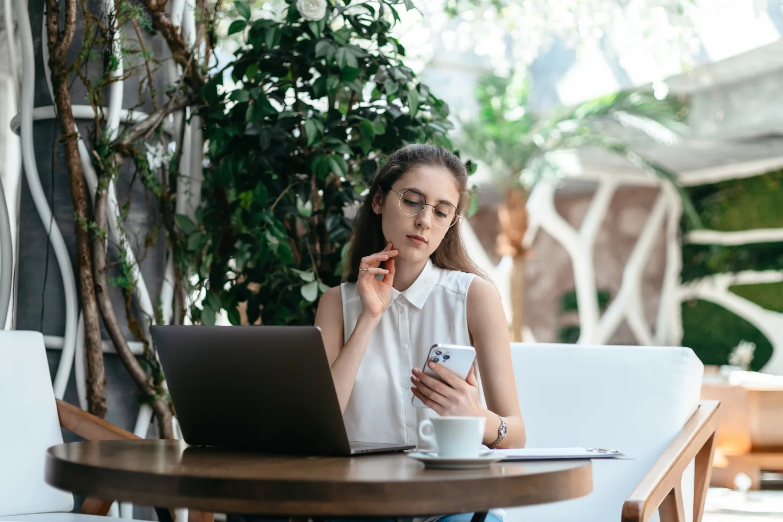A thoughtful business owner at a cafe table with her phone and laptop, weighing which assistant to pay for