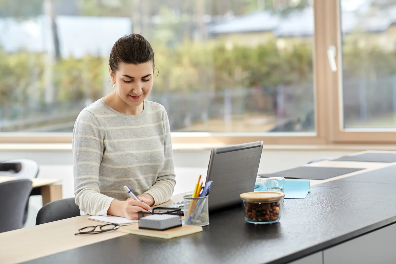 A business owner at her home office desk writes a note beside the laptop, the customer record in the middle of the day