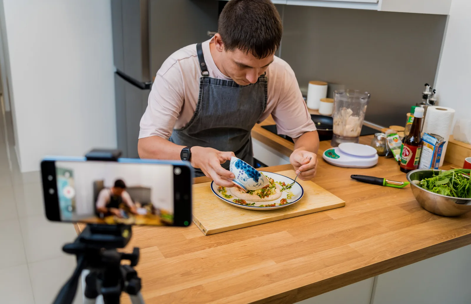 A cook plates a dish at the kitchen counter while a phone on a tripod films it