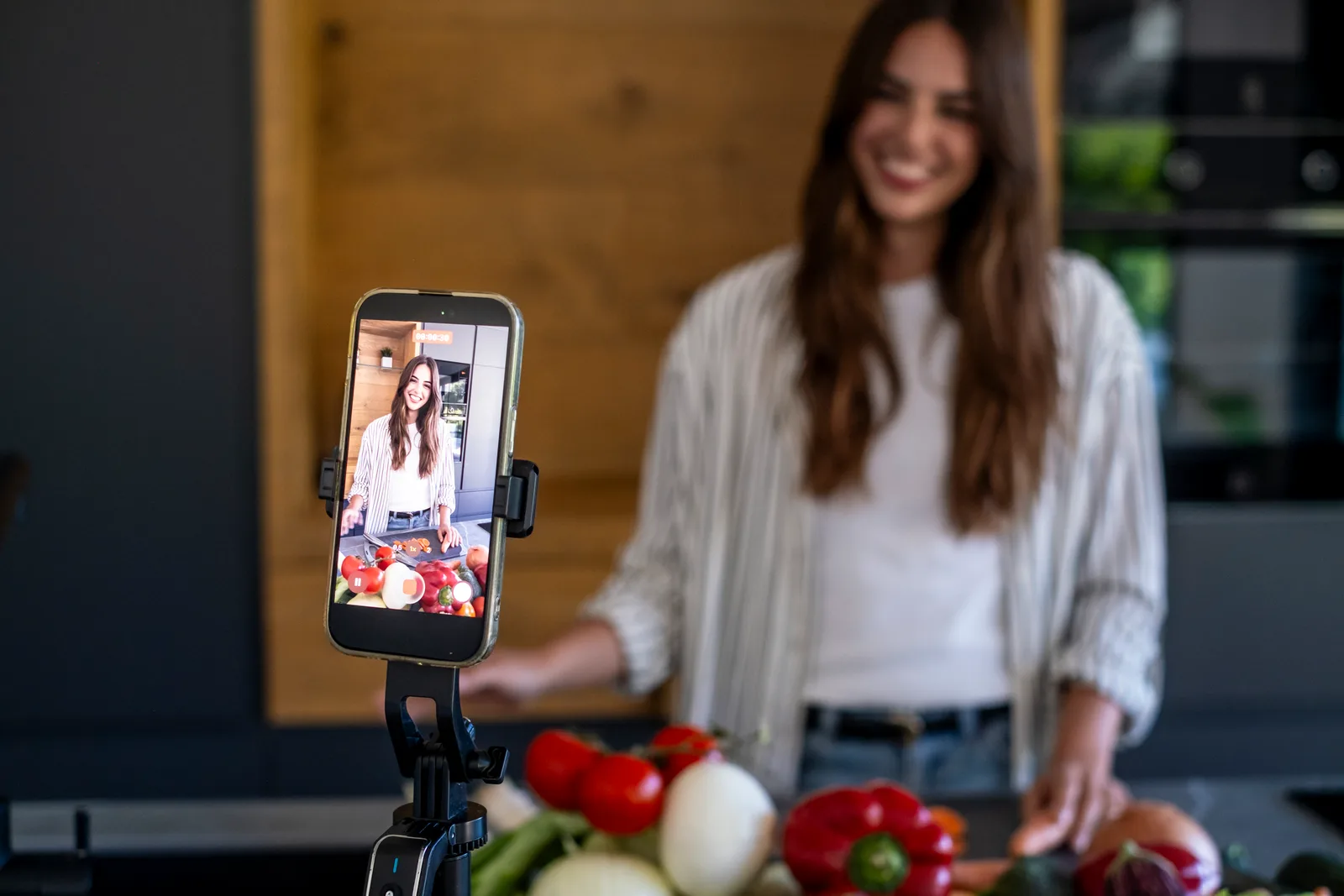 A smiling cook films a food video in her kitchen with a phone on a small tripod