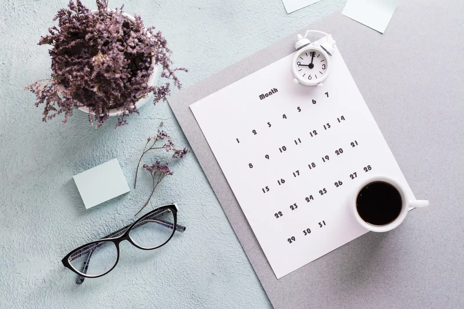 A blank monthly calendar sheet lies beside a small alarm clock, a cup of coffee and reading glasses