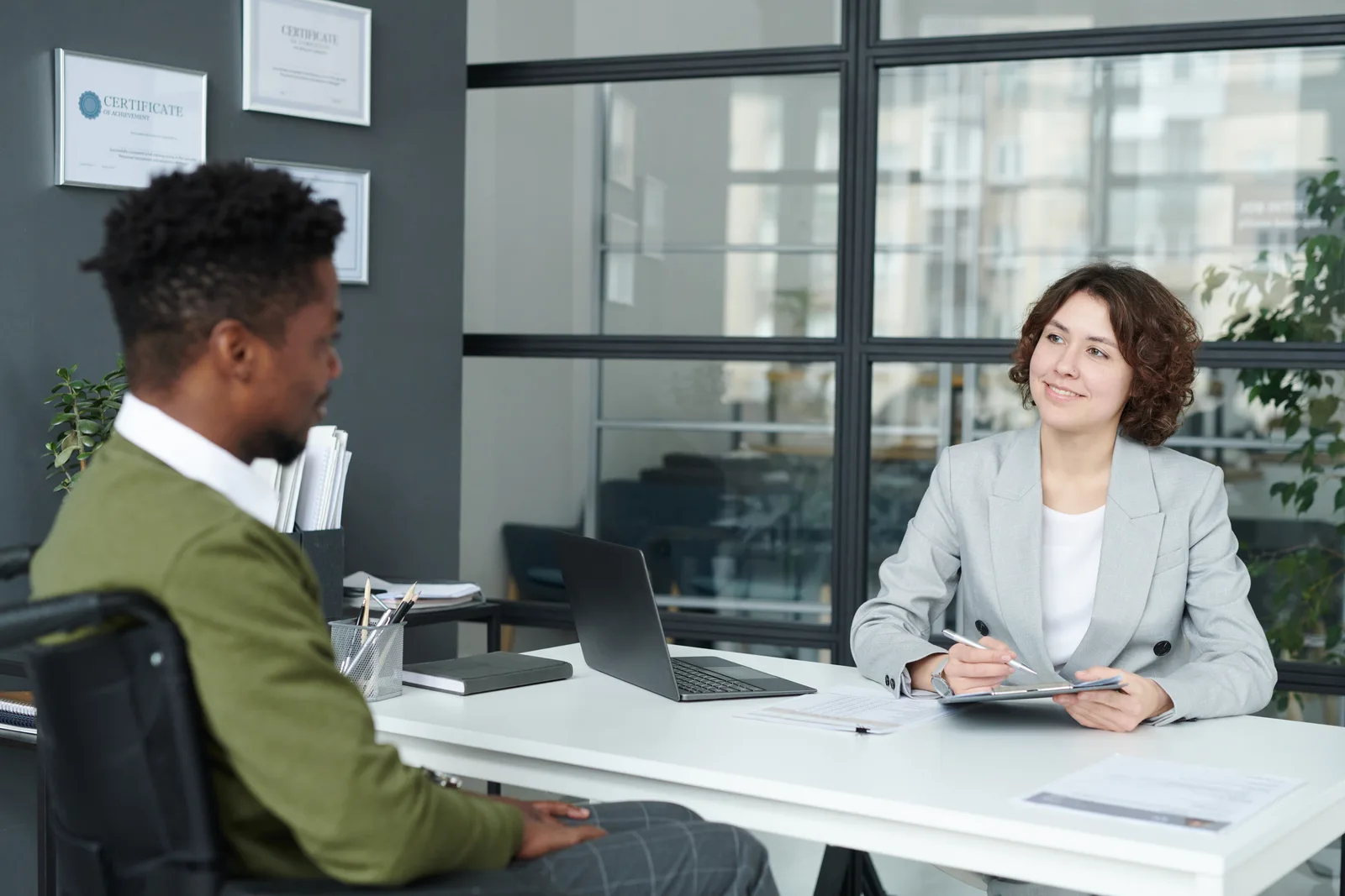 A hiring manager and a candidate talk across a desk in a small office, a laptop open between them