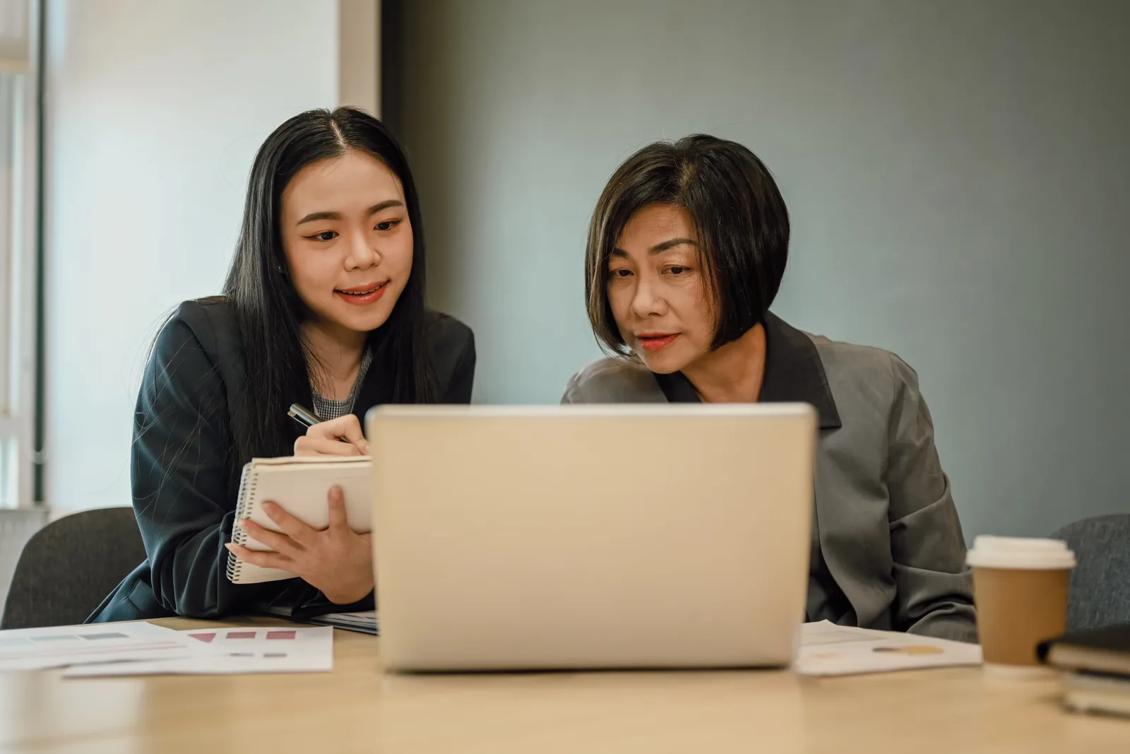 Two colleagues sit at a table comparing figures on a laptop screen, printed charts spread beside them