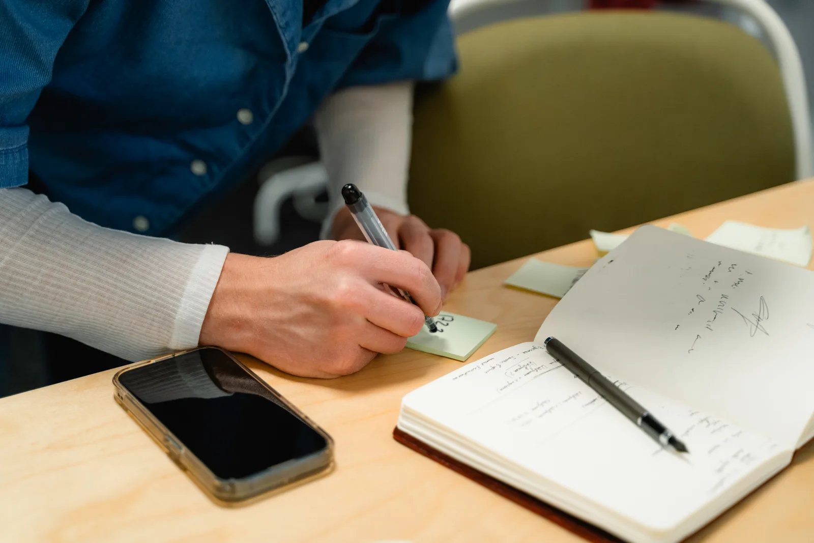 A person writes on a sticky note beside an open notebook, a phone and a pen on a wooden desk
