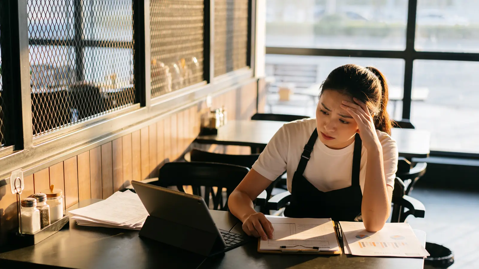 A cafe owner in an apron sits at a table with a laptop and paperwork, head resting on one hand