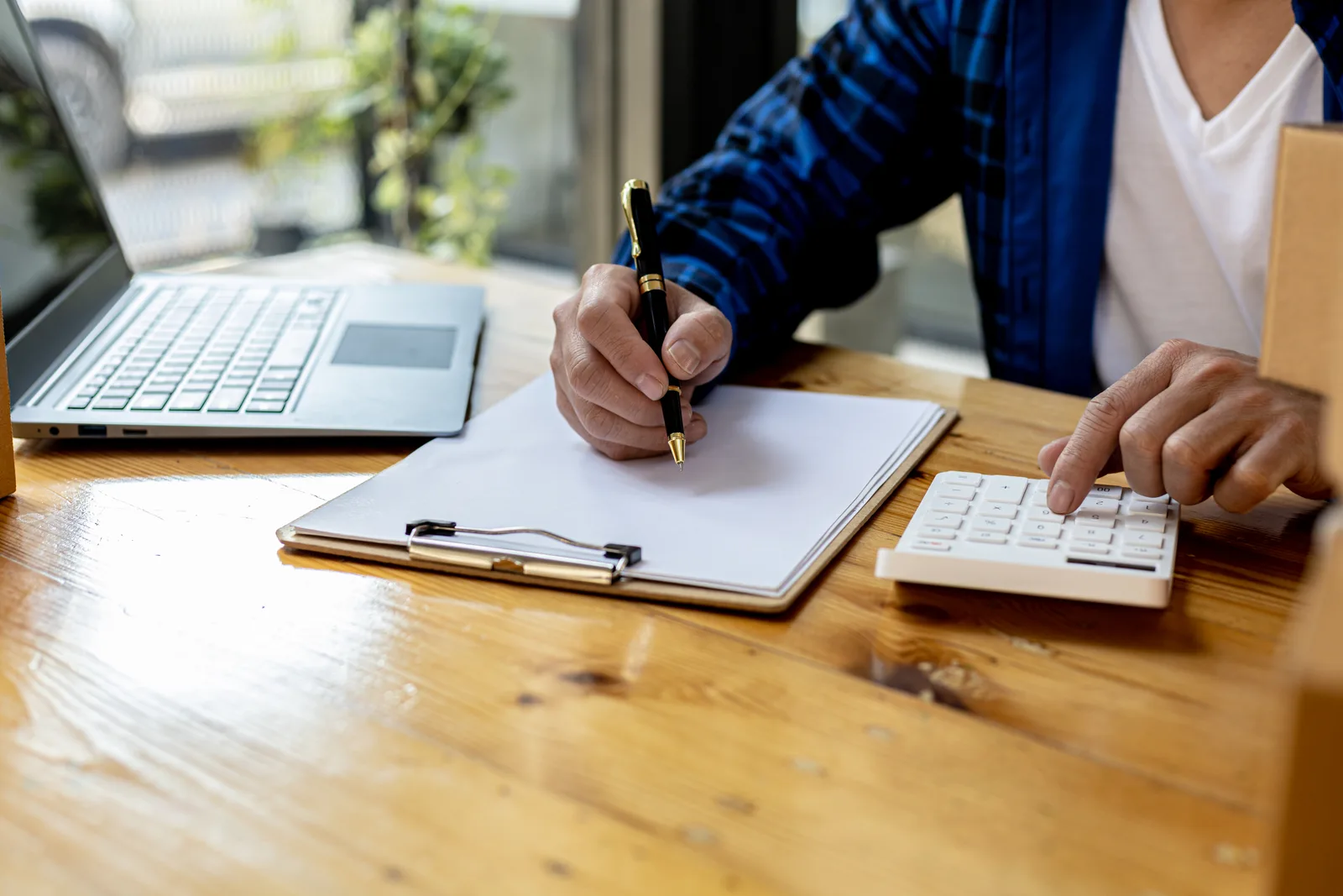 Someone signs a form on a clipboard beside a calculator and an open laptop on a wooden table