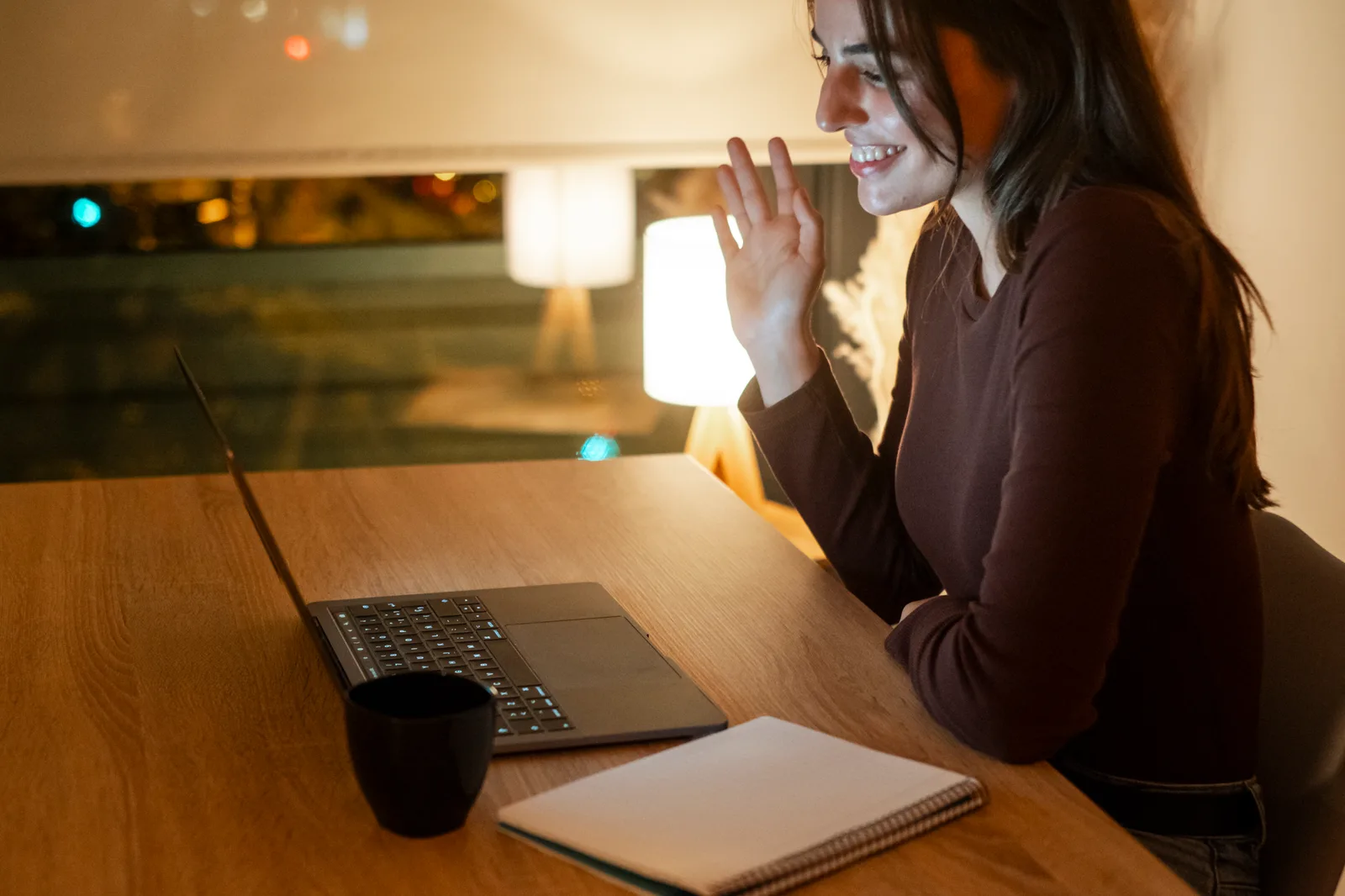 A remote worker waves on an evening video call at her laptop, a lamp lit and city lights in the dark window
