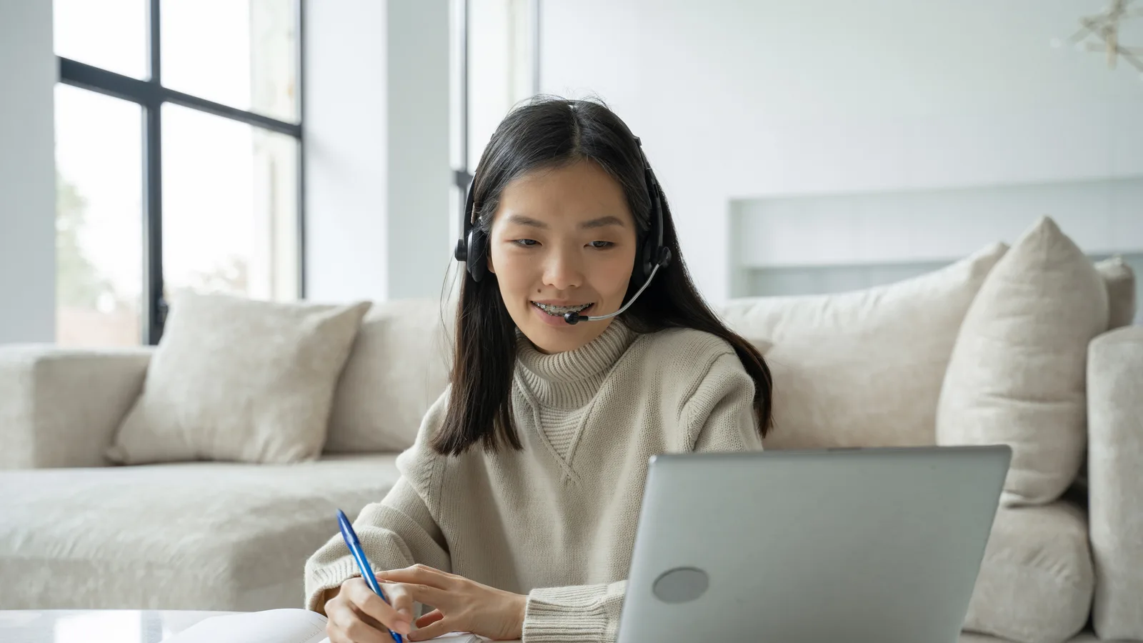 A virtual assistant in a headset with a microphone takes notes beside her laptop in a bright living room