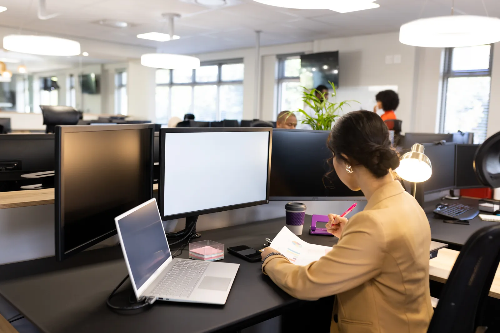 An assistant marks up a printed report at her open-plan office desk, with monitors and a laptop in front of her