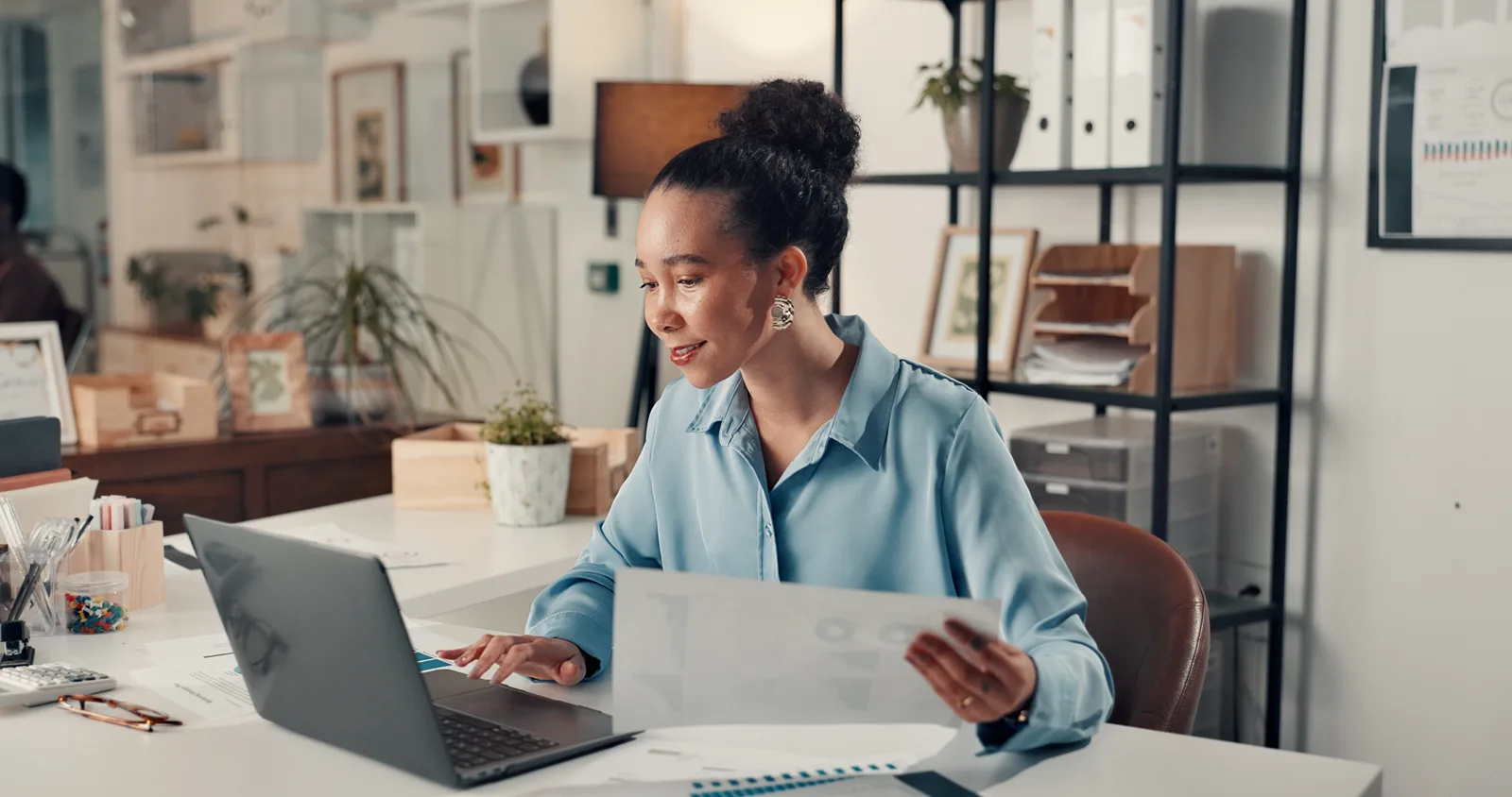 A woman types on her laptop at a small-office desk, a calculator and a printed chart report beside her