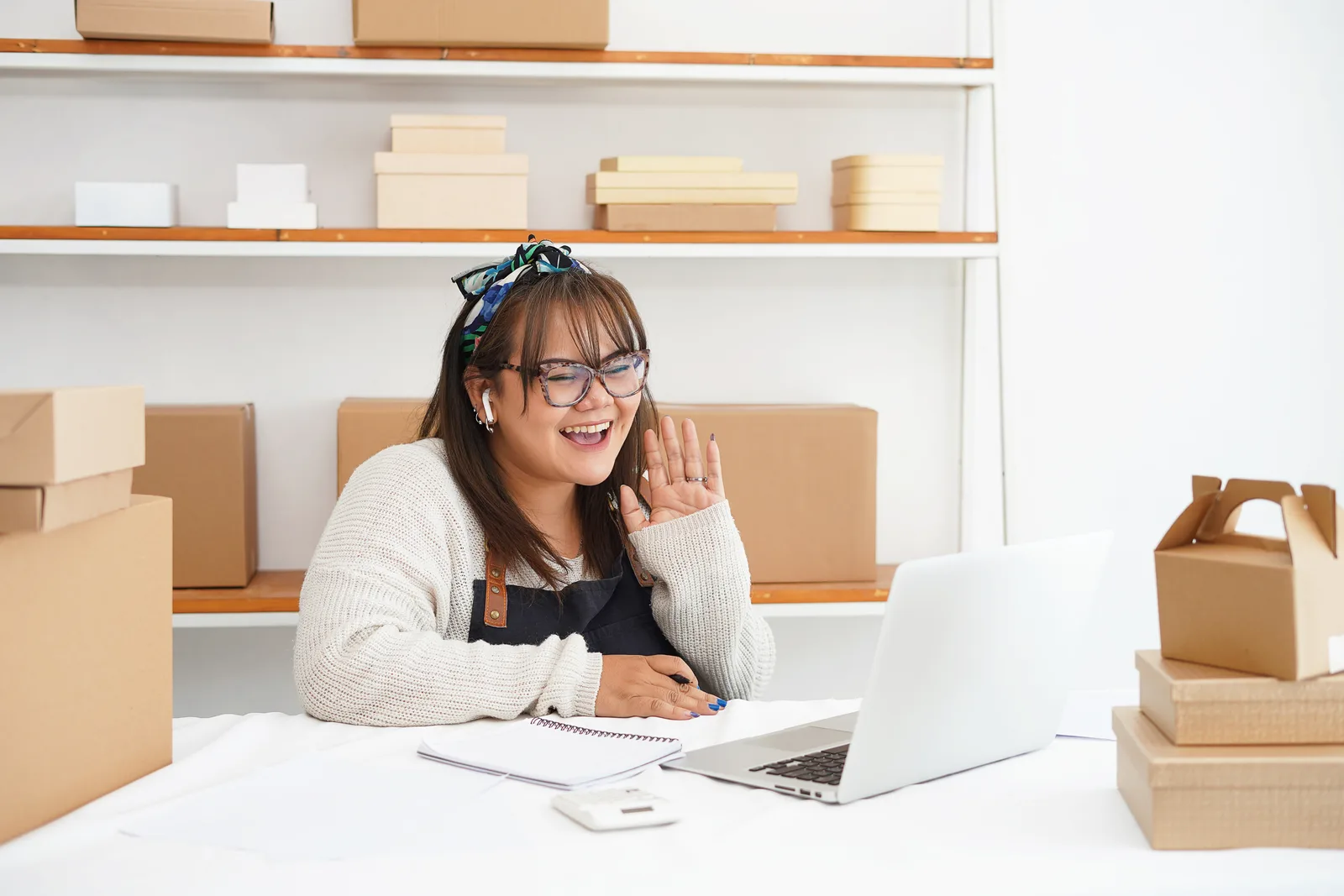 A shop owner in an apron waves hello on a laptop video call in her stockroom, a notebook open on the desk