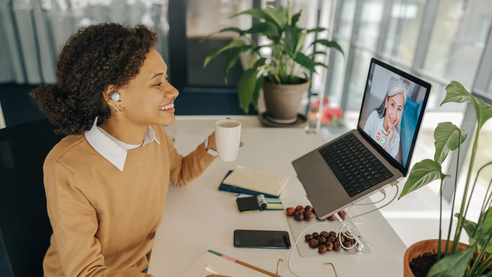 A smiling woman holds a coffee mug on a laptop video call with another woman, notebooks and a phone on her desk