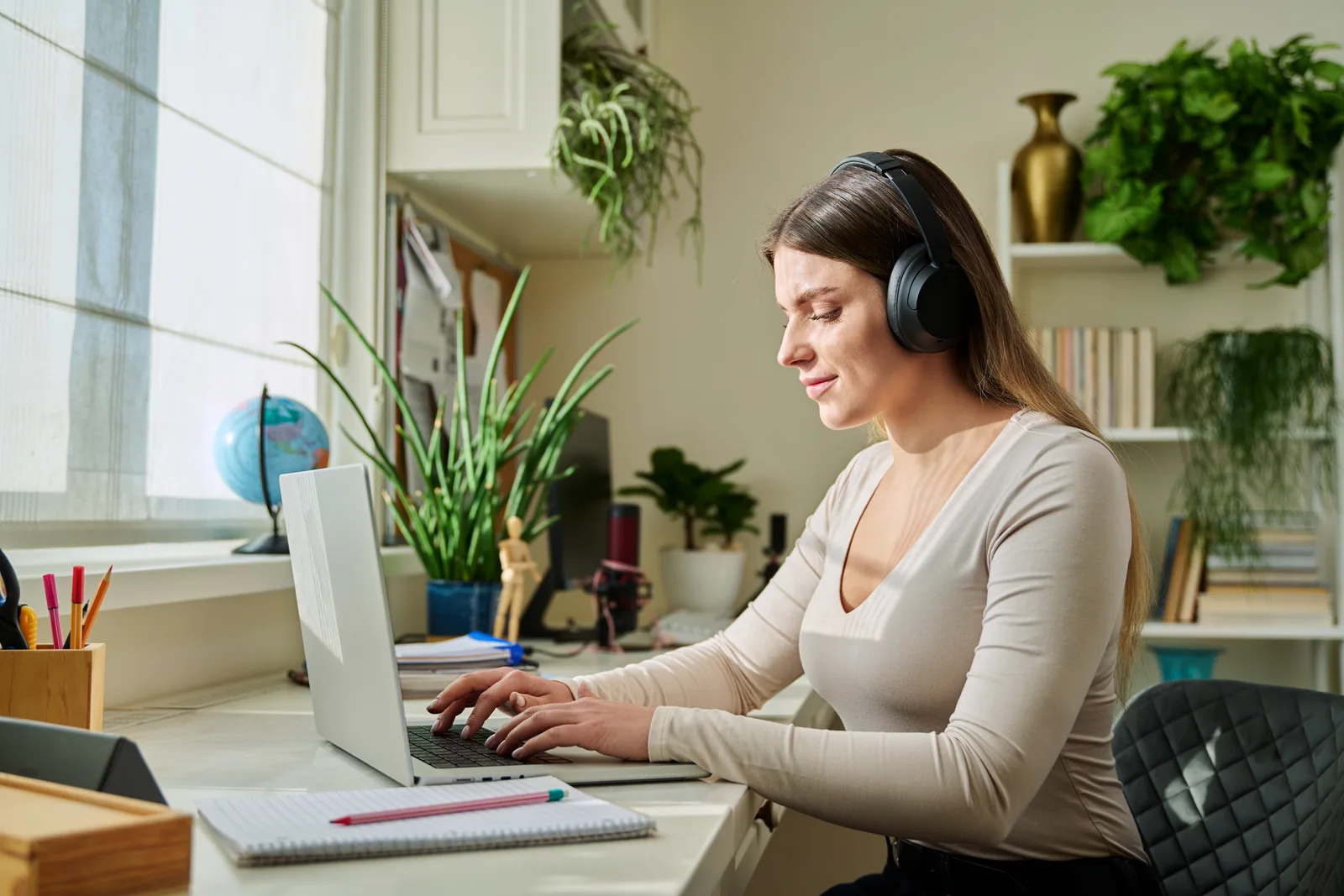 A woman in over-ear headphones types on a laptop at her home office desk, a notepad and pencil beside her
