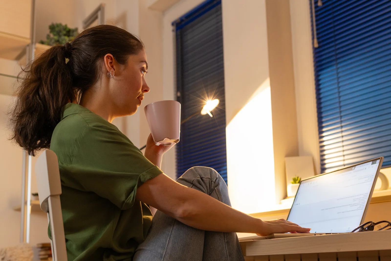 A woman sips coffee and works on her laptop at home as low sun slants through the window blinds