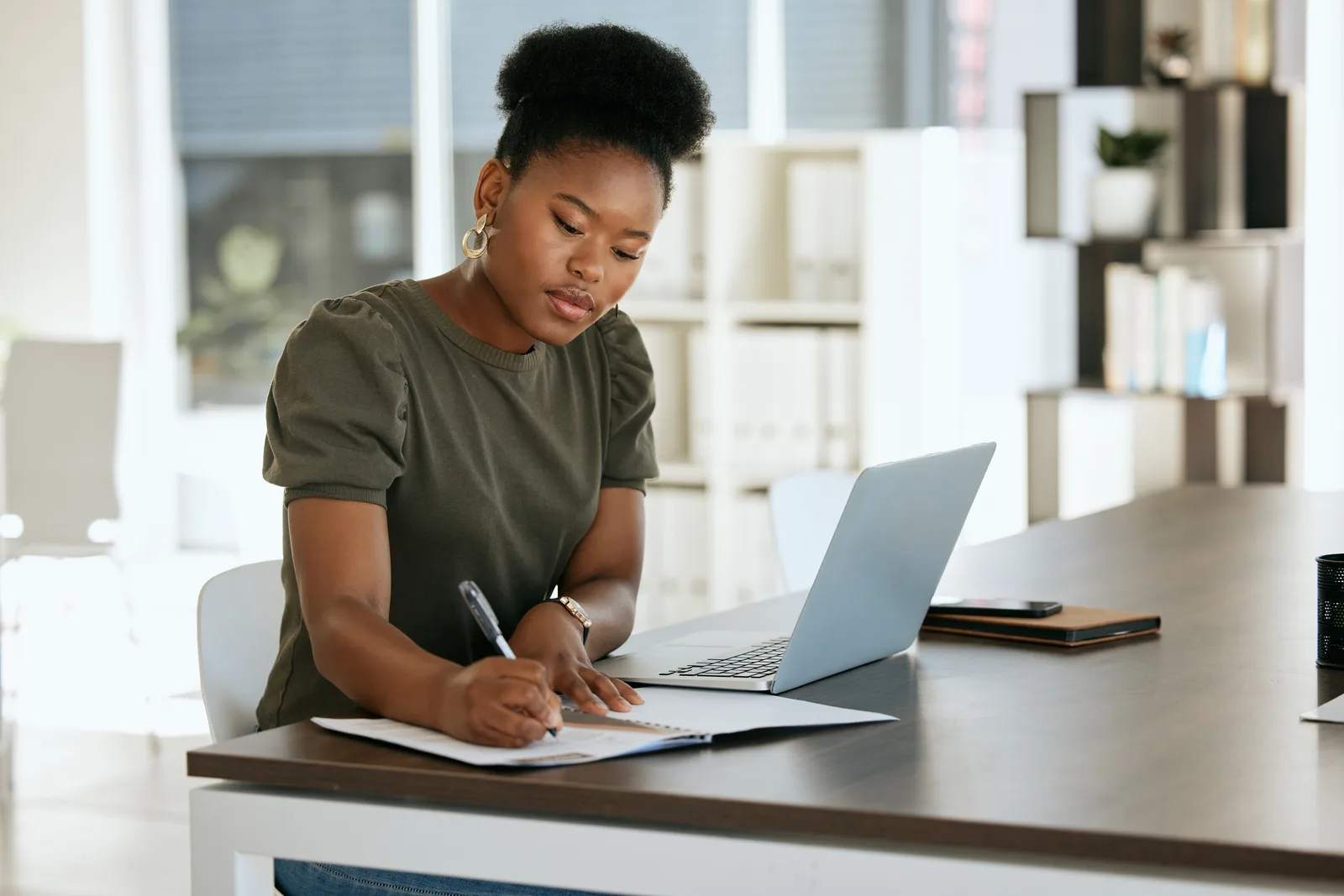 An executive assistant writes notes on a printed page beside her open laptop at a tidy desk in a bright office
