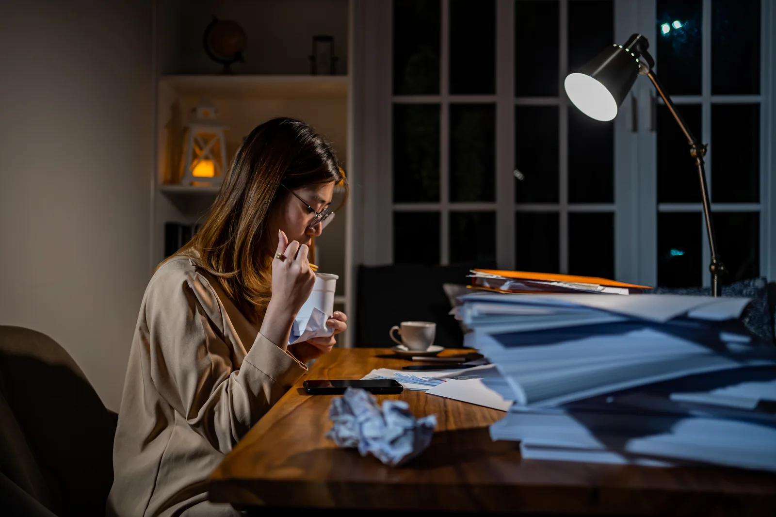 A business owner works late by lamplight at her home desk, tall stacks of paperwork and a crumpled page beside her