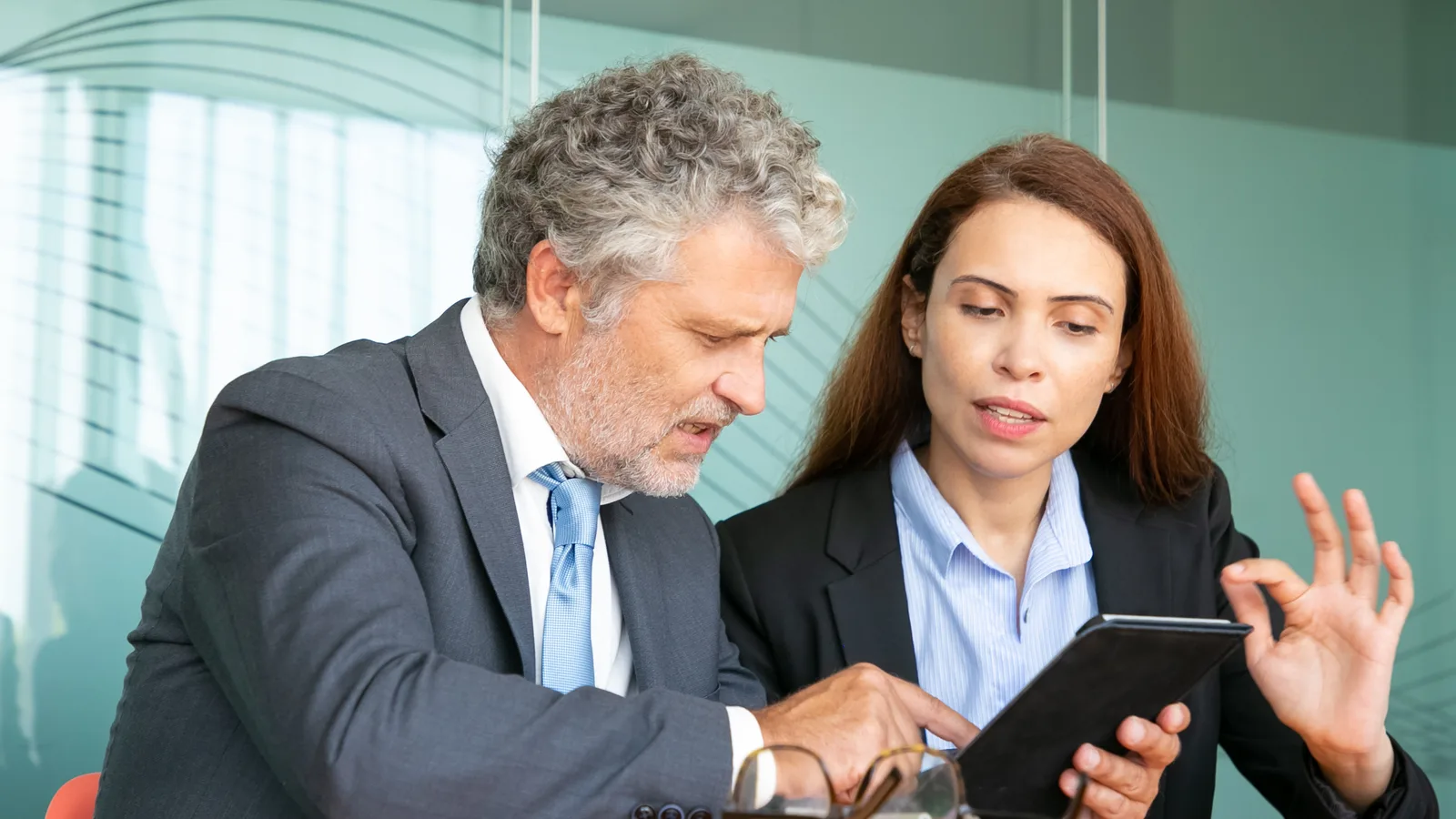 An executive assistant holds up a tablet while the gray-haired executive beside her points at the screen in a glass office