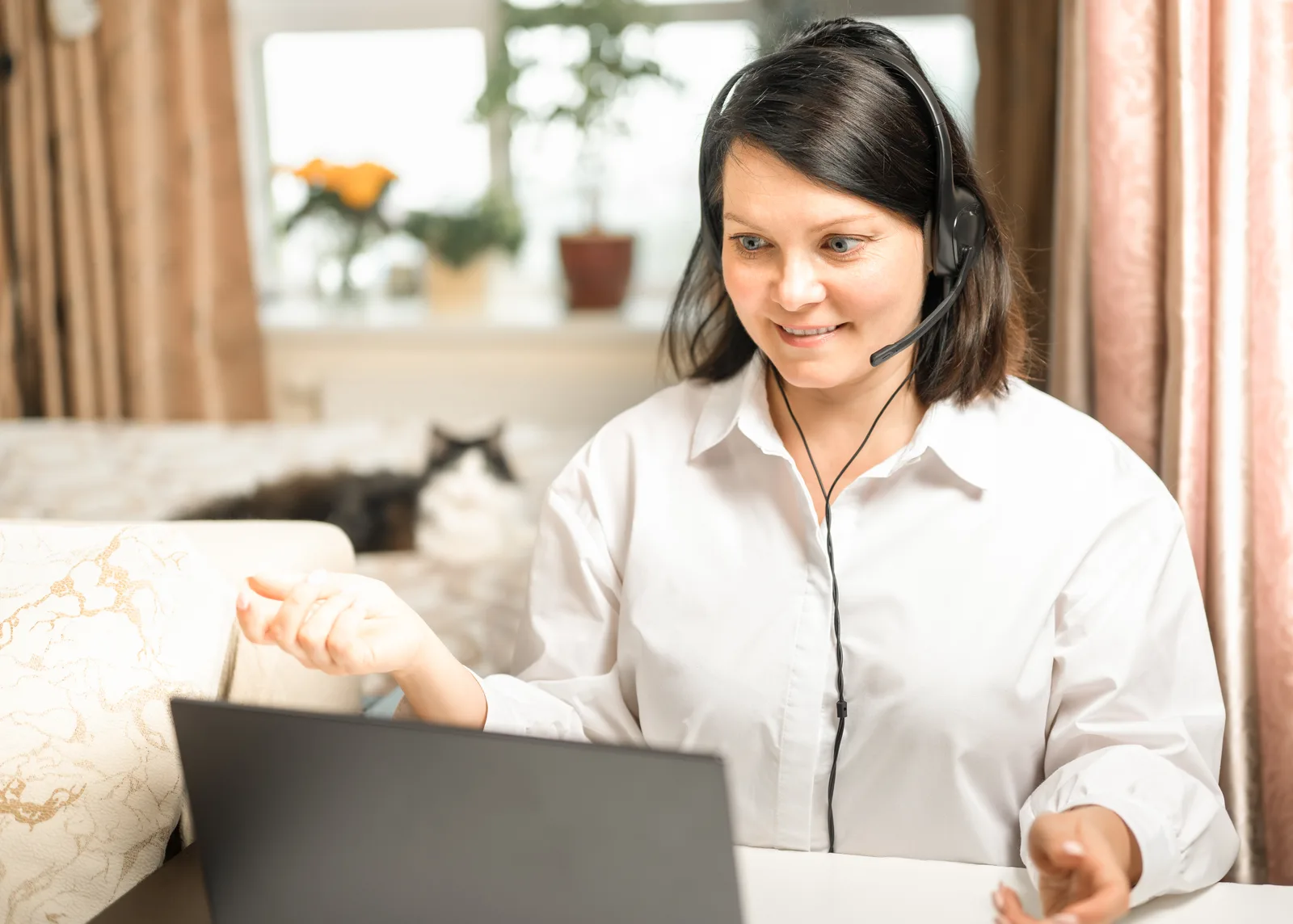 A remote executive assistant in a headset talks to her laptop screen from her living room, a cat on the sofa behind her