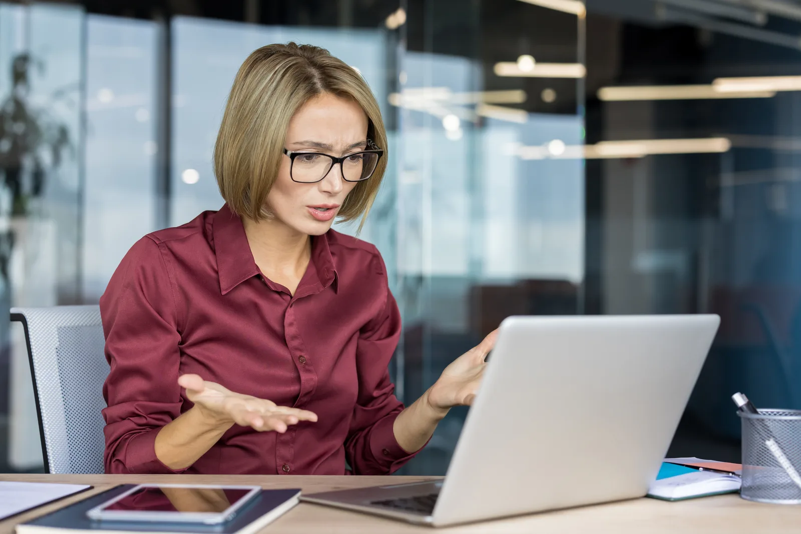 A woman in a red shirt frowns at her laptop screen and raises both hands, puzzled, at an office desk
