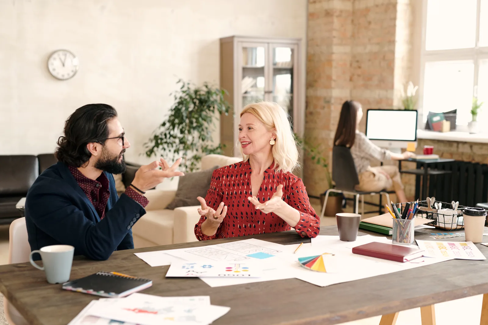A man and a woman talk across a table spread with printed charts in a brick-walled studio office