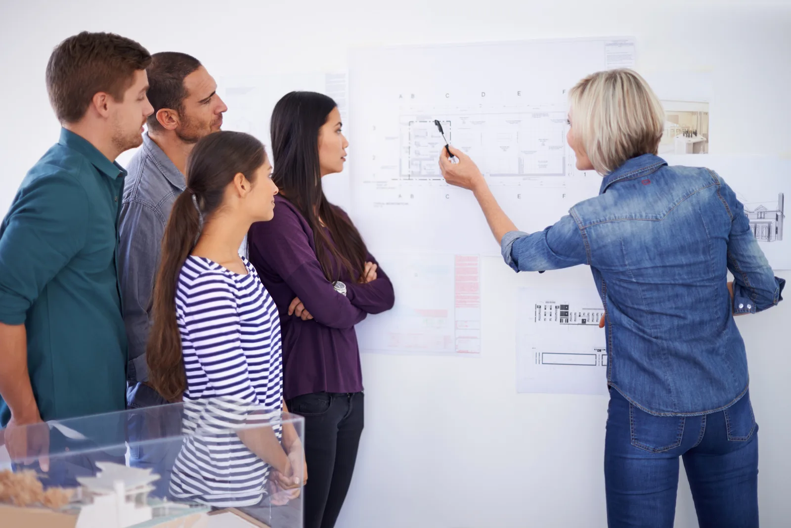Five colleagues stand at a wall of printed plans while one of them points to a section with a marker