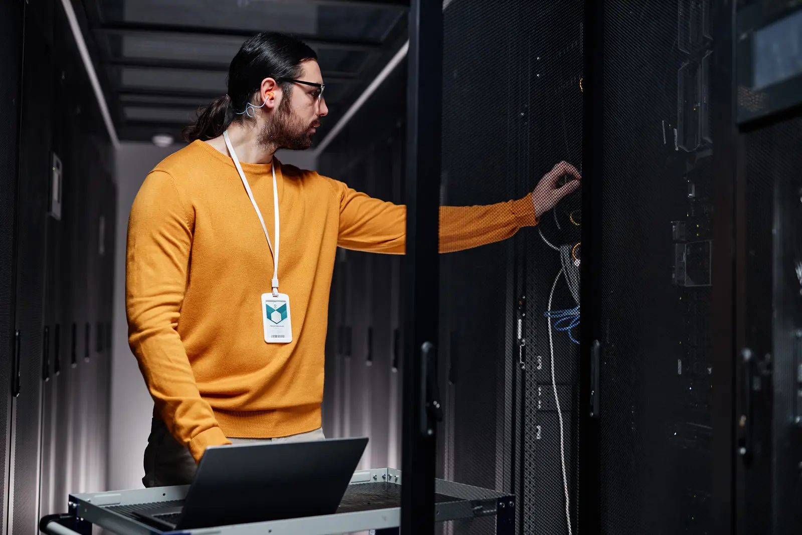 An engineer in an orange sweater reaches into a server cabinet, a laptop open on a cart beside him